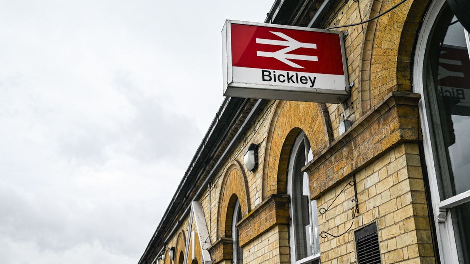 A close-up view of Brockley train station's exterior with a prominent red and white National Rail sign displaying 'Bickley' for the station name, attached to a yellow brick building featuring arched w