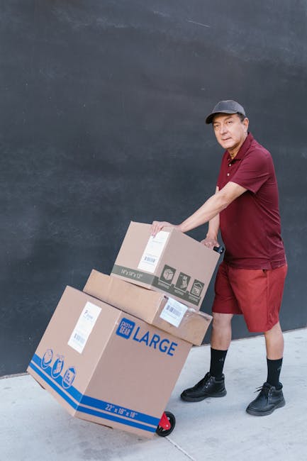 A delivery person dressed in a maroon polo shirt, matching shorts, and a grey cap is pushing a hand truck loaded with three cardboard boxes, one labeled 'LARGE' and another with shipping labels, again