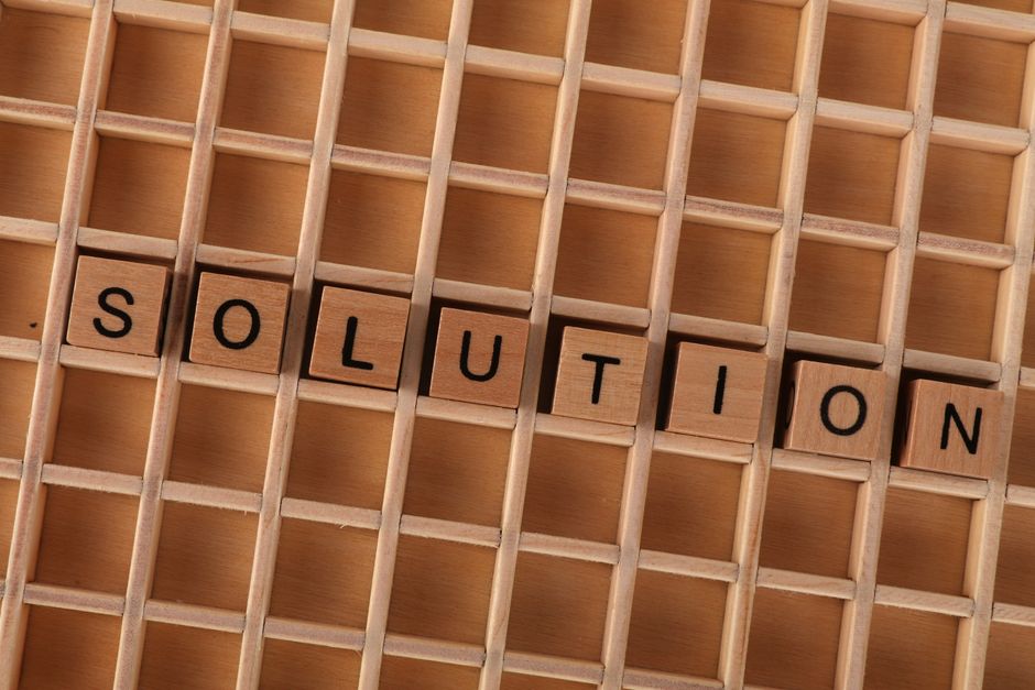 A close-up view of a wooden grid display with small square compartments, on which the word 'SOLUTION' is spelled out using black letter tiles. The background features smooth, light-colored wood, highl