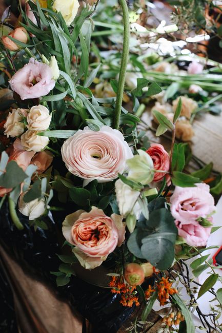 A close-up view of a floral arrangement featuring a variety of fresh flowers, including large, pale pink ranunculus with tightly layered petals, soft peach and blush lisianthus, and delicate orange an