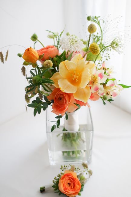 A vibrant floral arrangement featuring large, soft yellow and peach roses, delicate pink and white alstroemeria, and small, rounded craspedia flowers, all set in a clear glass vase. The bouquet includ