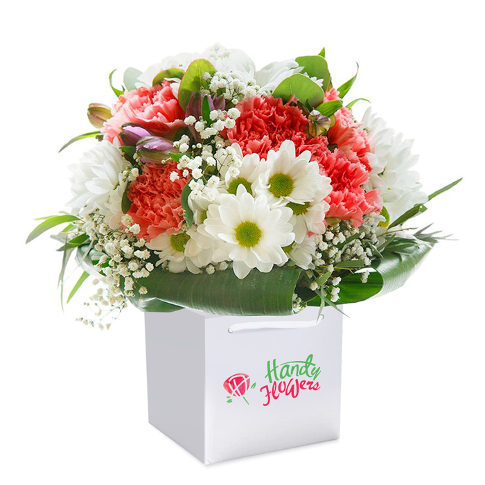 Standing tall within a modern white gift bag, this meticulously arranged bouquet showcases a dynamic interplay of colours and textures, reminiscent of a stroll through Brockley's vibrant neighbourhood gardens. At the bouquet's centre, frilled coral and crimson carnations burst with lively energy, each petal slightly tousled yet artfully arranged for optimal fullness. Interwoven throughout, clusters of crisp white daisies with vivid lime-green centres evoke a sense of openness and calm, their simple beauty balanced by the ethereal lightness of scattered white baby's breath. Accents of unopened flower buds edged in soft purple nestle subtly on the arrangement's edge, contributing to an elegant sense of anticipation and surprise-echoing the hidden corners and diverse plantings seen around Brockley's Conservation Area. Enveloping the florals, a rich mantle of deep green, broad-leaved foliage shapes the bouquet into a harmonious, rounded whole, the sheen of the greenery forming a visually stunning backdrop. The bouquet sits illuminated by gentle, natural light on a seamless white background, highlighting the freshness and ready-to-gift composition. Complete with sturdy white ribbon handles for effortless delivery, this refined arrangement is perfectly suited for those special Brockley moments-be it a birthday, anniversary, or spontaneous celebration-reflecting both the neighbourhood's creative spirit and the florist's expert touch.