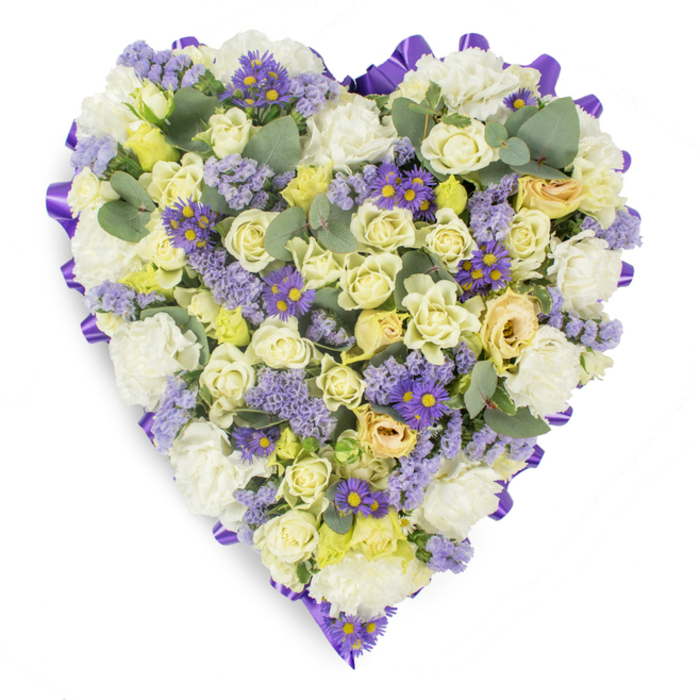 A meticulously constructed heart-shaped sympathy arrangement pictured on a pristine white background, the design reads as an intimate study in soft colour and refined texture. Cream and pale yellow roses dominate the surface, their petals ranging from tight, sculpted buds to gently opening blossoms that offer a layered, luxurious tactile quality; these are grounded on a cushion of ruffled white carnations that add fullness and a classical, consoling presence. Interspersed among the neutrals are delicate sprays of lavender statice and vivid purple aster-like blooms with bright yellow centres-small, joyful accents that lend depth and emotional contrast without overwhelming the arrangement's serenity. Blue-green eucalyptus leaves, rounded and matte, are artfully positioned to punctuate the composition, introducing silvery highlights and a cooling, herbal scent implied by their visual texture. The whole heart is edged with a continuous rich purple satin ribbon, neatly looped to create a formal, polished border that punctuates the silhouette and elevates the piece for ceremonial use. Photographed with soft, diffused lighting, every petal and ribbon fold is rendered with clarity, reflecting careful floristry technique. Arranged by Florist Brockley for families in Brockley and surrounding neighbourhoods-whether for a church service near St John's, a quiet graveside at Ladywell, or a wake close to Brockley Station-this tribute conveys steady love, respect, and consolation, making it a thoughtful, traditional choice for honouring a loved one.