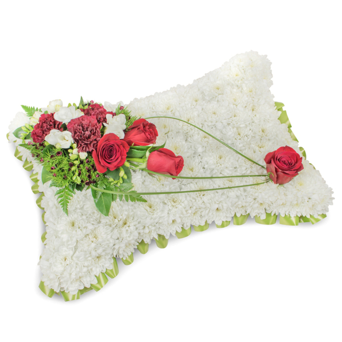 An elegant sympathy cushion, shaped as a gentle rectangle, its surface completely covered with hundreds of small white chrysanthemums so closely placed that the form reads like a soft, white carpet. The texture is plush and uniform, reflecting light in a way that emphasises the pillow's peaceful presence; a narrow lime-green ribbon or foliage border lifts the edge and provides a fresh, handmade finish. At the upper left an abundant cluster of deep red roses and dusky burgundy carnations creates the arrangement's focal point-roses in varied opening stages, their velvety petals contrasting vividly against the snow-white chrysanthemum ground. Interspersed among these are tiny white freesias or fine spray chrysanthemums that brighten the composition and introduce delicate vertical accents. The greenery includes feathery fern fronds and glossy leaves that give depth and a gardener's lushness to the piece. From this primary spray, three slender, arching stems of bear-grass or similar vivid green foliage trace a graceful diagonal toward the lower right quadrant, culminating in a single prominent red rose that appears cradled by the sweeping lines-a deliberate touch signifying remembrance held in tender hands. The overall impression is tranquil and dignified; the scent would be subtle, a gentle mingling of white chrysanthemum cleanness, faint freesia sweetness and green leaf. Suited for a funeral service, memorial or graveside tribute in Brockley and surrounding neighbourhoods such as New Cross or Ladywell, this pillow balances craftsmanship with heartfelt sentiment, a classic floral language for saying goodbye with respect and love.