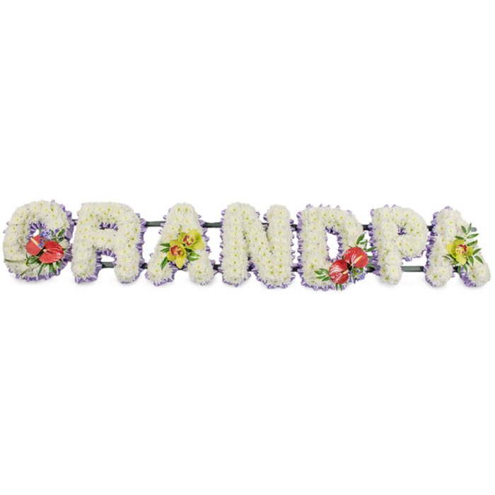 This image shows a thoughtfully crafted funeral flower tribute spelling out the word GRANDPA in large, upright block letters laid in a long row, ready to rest on trestles at a church or graveside in Brockley. Each individual letter is filled edge to edge with compact white chrysanthemums, their small, multi-petalled blooms packed closely together to create a dense, velvety cushion of pure white. The surface catches the light softly, giving the entire tribute a gentle glow against the plain white background. At the base of each letter runs a subtle trim of pale lilac ribbon or fabric, peeping out just enough to add a hint of cool colour and to frame the shapes so they read clearly from a distance, whether set near Hilly Fields or outside a local funeral home. Behind the flowers, dark green framework links the letters, barely visible but essential for supporting the weight of the design. Touches of richer colour are woven into specific points. On the far left, the letter 'G' is adorned with a cluster of deep red anthuriums, their high-shine, heart-shaped petals and bright central spadices surrounded by fresh green leaves and perhaps tiny flecks of purple filler, creating a focal moment of passion and devotion. The first 'A' carries a more delicate spray of yellow-green orchids, their starry petals and softly arching stems nestled into foliage, lending refinement and a sense of enduring beauty. In the middle, the letter 'D' repeats the bold red anthuriums, while the final 'A' mirrors the earlier orchids, providing visual and emotional symmetry. Overall, the piece feels dignified and deeply affectionate, a personalised floral tribute that conveys love, gratitude, and remembrance for a grandfather cherished by family in Brockley and nearby streets.